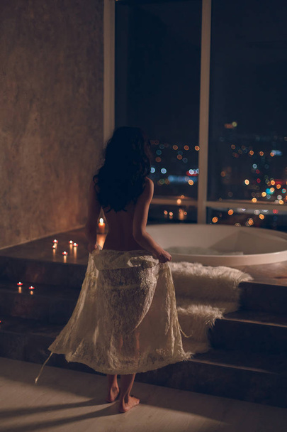 Sensual young woman staying naked or nude with white lace robe on her hips getting ready to take a bath in luxury hotel room at night with awesome city lights view trough big window. - Foto, afbeelding