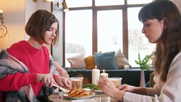 Duas jovens sentadas no café e conversando enquanto comem panquecas. Grande interior moderno
 - Filmagem, Vídeo