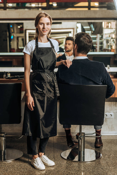 bella giovane parrucchiere sorridente alla macchina fotografica mentre il cliente maschio seduto in sedia al salone di bellezza
  - Foto, immagini