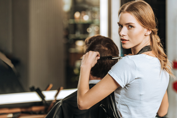 schöne junge Friseurin schaut in die Kamera, während sie dem männlichen Kunden im Schönheitssalon die Haare kämmt und schneidet  - Foto, Bild