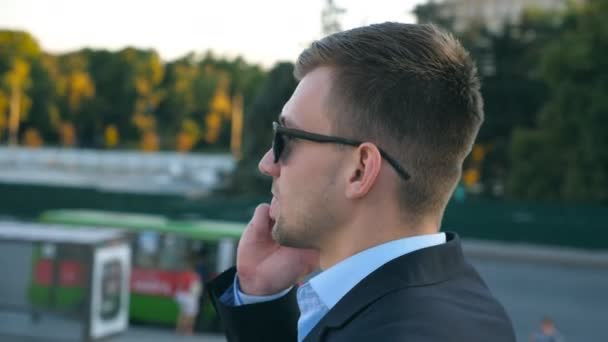 Camera spinning around of handsome businessman talking on phone outdoor. Young business man in sunglasses having conversation on cellphone. Portrait of guy speaking on smartphone outside. Slow motion - Materiał filmowy, wideo