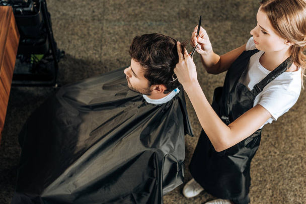 Hochwinkelblick des Friseurs, der einem hübschen jungen Mann im Schönheitssalon Haare schneidet  - Foto, Bild
