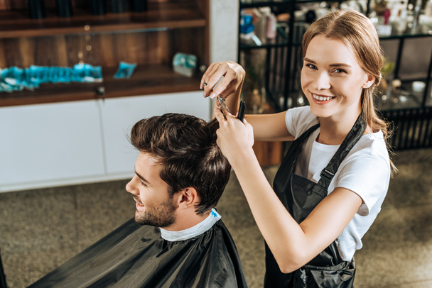 vista de ángulo alto del estilista sonriendo a la cámara mientras corta el cabello a un cliente guapo en el salón de belleza
 - Foto, Imagen