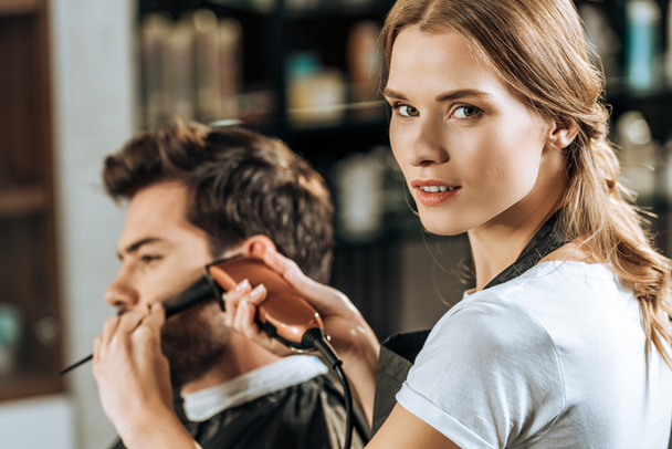 ελκυστική hairstylist βλέπουν φωτογραφική μηχανή, ενώ το ψαλίδισμα των τριχών σε άνδρες πελάτης στο σαλόνι ομορφιάς  - Φωτογραφία, εικόνα