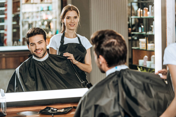 улыбающийся молодой парикмахер и красивый клиент, смотрящий на зеркало в салоне красоты
 - Фото, изображение