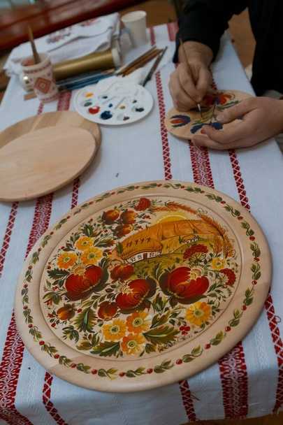 Petrykivka estilo de pintura placa de madeira, paisagem retro ucraniana típica, mãos do homem handpainting na placa redonda de madeira decorativa artesanal
 - Foto, Imagem
