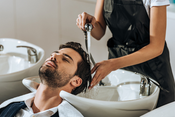 cropped shot of hairstylist washing hair to handsome young man in beauty salon - Foto, Bild