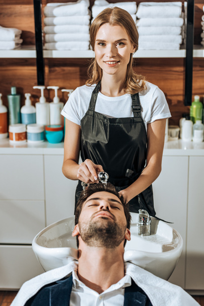 beautiful hairdresser smiling at camera while washing hair to handsome man in beauty salon - Фото, зображення