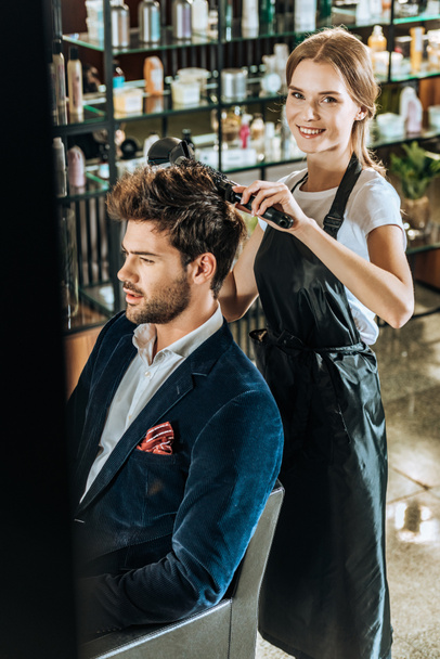 young female hairdresser smiling at camera while drying hair to handsome client in beauty salon - Foto, Imagem