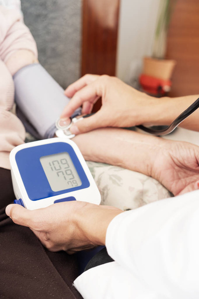 close-up de um homem médico caucasiano, em um casaco branco, medir a pressão arterial de uma mulher paciente caucasiana sênior com um esfigmomanômetro, em sua casa ou em um lar de idosos
 - Foto, Imagem