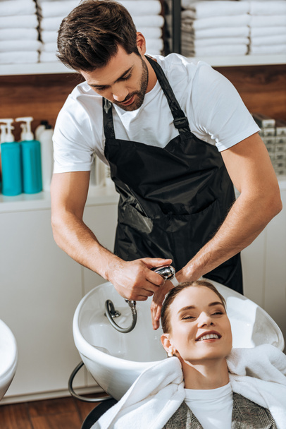 handsome young hairdresser washing hair to smiling young woman in beauty salon - Photo, Image