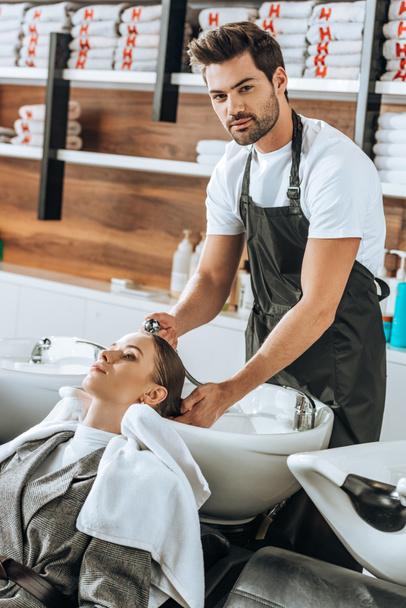 guapo joven peluquero lavando el cabello a la mujer y mirando a la cámara en el salón de belleza
  - Foto, Imagen