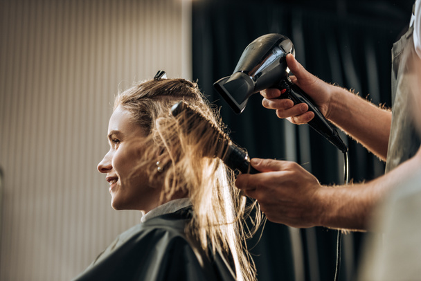 Oříznout záběr kadeřník sušení vlasů na usmívající se dívka v salonu krásy   - Fotografie, Obrázek