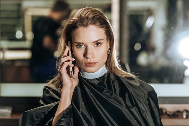 attractive young woman talking by smartphone and looking at camera in beauty salon - Foto, afbeelding