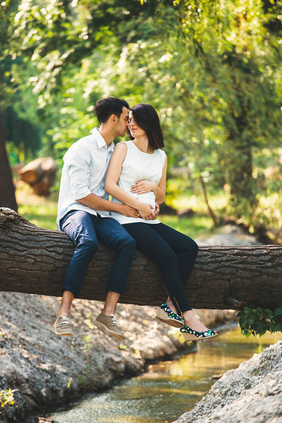 Beautiful pregnant couple relaxing outside in the forest, while sitting and kissing on a tree stem above a rivulet with sun light penetrating through green leaves in the background. - Photo, Image