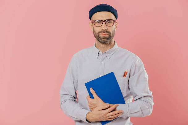 Shot of serious unshaven young European male looks confidently at camera, has dark bristle, holds notepad for noting records, dressed in formal shirt and headgear, models against pink background. - Photo, Image