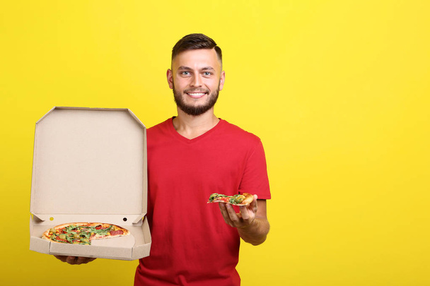 Young man eating pizza and holding cardboard box on yellow background - Valokuva, kuva