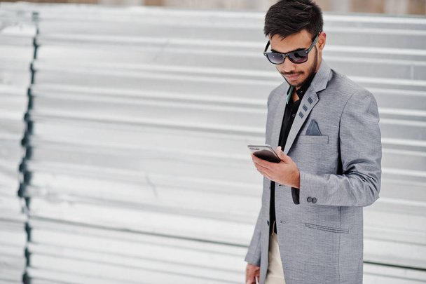 Casual young indian man in silver jacket and sunglasses posed against steel background, looking at mobile phone. - Photo, image