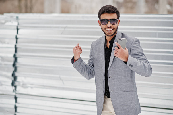 Casual young indian man in silver jacket and sunglasses posed against steel background, showing to mobile phone. - Foto, imagen