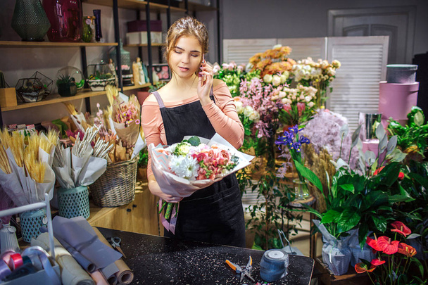 Busy young florist hold bouquet in hand. She talk on phone. Young woman stand in room full of flowers and plants. - Valokuva, kuva