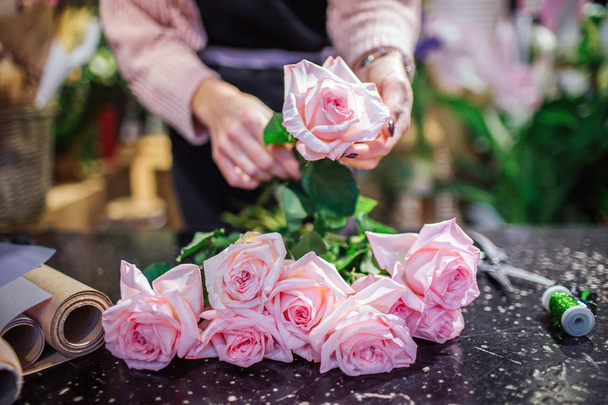 Close of beautfiul light pink roses lying on table. Florist hold one flower in hand. Paper rolld and coil with green thread are on table. - Foto, imagen