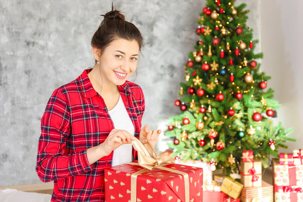 Young beautiful brunette woman in plaid checkered pajamas in decorated bedroom interior with gift box and Chrictmas tree on background. People on Christmas morning concept. Close up, copy space. - Foto, Imagen