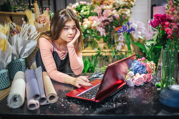 Amazed young florist look at red laptop and wonder. She touch keyboard and lean on table. Paper rolls are there as well. Plants behind her. - Fotografie, Obrázek