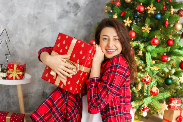 Young beautiful brunette woman in plaid checkered pajamas in decorated bedroom interior with gift box and Chrictmas tree on background. People on Christmas morning concept. Close up, copy space. - Fotoğraf, Görsel