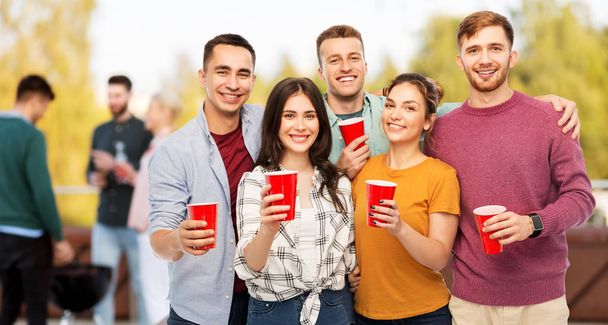 celebration, friendship and people concept - group of smiling friends toasting non alcoholic drinks in party cups over rooftop party background - Фото, изображение