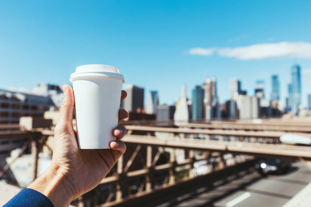 częściowy widok mężczyzny trzymającego jednorazowe filiżankę kawy z niewyraźne Nowy Jork na tle - Zdjęcie, obraz