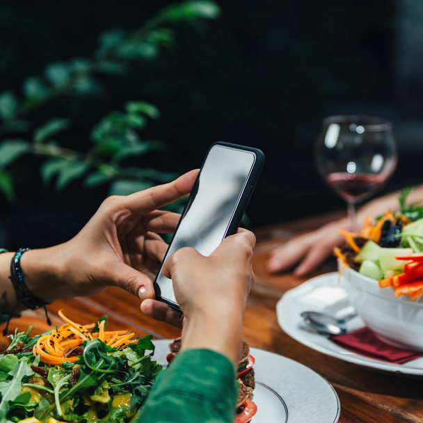 Square image of young woman photographing delicious vegetarian burger and mixed organic salad on white plate in vegetarian restaurant. - Photo, Image
