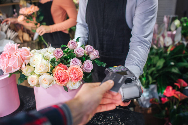 Cur view of man holding credit card in hand. He give it to florist. Flower seller has thereminal for cards. Table ull of colorful flowers. - Foto, Imagem