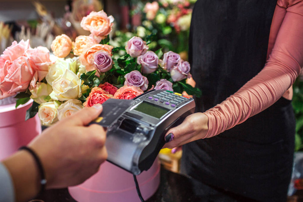 Cut view of man holding credit card above money therminal. He pays for flowers. Young female florist hold money therminal. Lots of flowers are behind her. - Photo, image