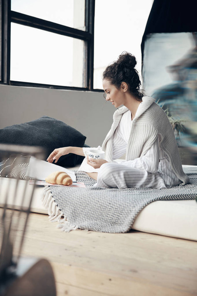 Cozy home portrait of young girl having breakfast on bed and reading magazine. Scandinavian room. - Fotografie, Obrázek
