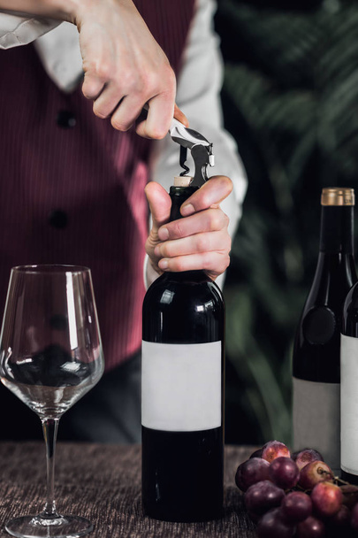 Жінка сомельє відкриває пляшку вина зі шпорою. Свіжий виноград і порожній келих вина на столі
.  - Фото, зображення