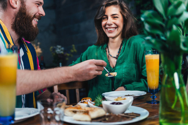 Happy couple enjoying romantic vegetarian dinner, drinking fresh orange juice on a date. Couple sitting at wooden table, wearing colorful outfit.    - Фото, зображення