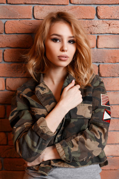 Portret van een charmante, sexy jongedame met mooie ogen in een trendy militaire camouflage groene jas en een grijs t-shirt in de buurt van de bakstenen muur. Krullend stijlvolle blond meisje. Moderne mode - Foto, afbeelding
