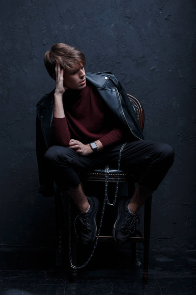 Стильный молодой человек в черном винтажном пиджаке в красном теплом гольфе в стильных брюках в модных кроссовках с металлической цепью сидит на деревянном стуле в комнате рядом со стеной. Привлекательный современный парень
 - Фото, изображение