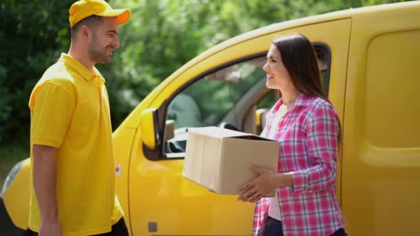 Delivery guy in yellow uniform gives parcel to female customer near car - Кадры, видео