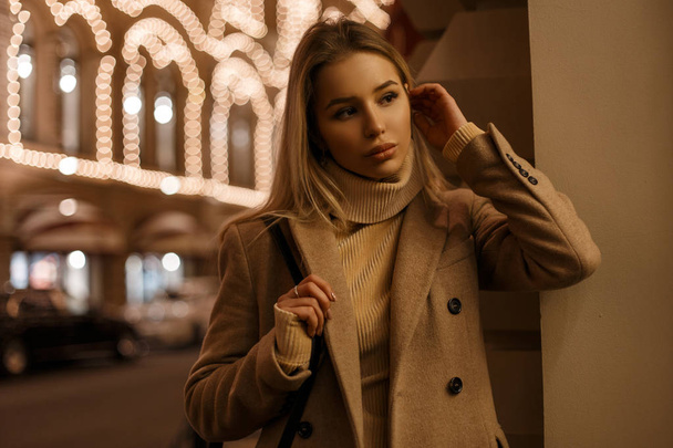 Удивительная красивая милая молодая женщина в модном элегантном теплом осеннем пальто и вязаном бежевом свитере моды, позирующая в ночном городе возле праздничных огней. Стильная очаровательная девушка
 - Фото, изображение