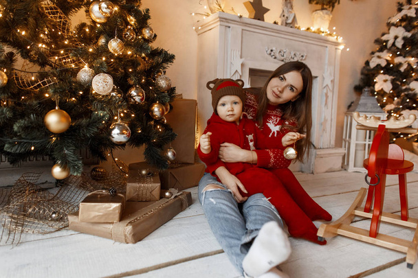 Fiatal vidám nő anya vintage vörös pulóverben ül a fa padló a stúdióban, és tartja a karjában a gyermeket a stílusos karácsonyi plüss ruhát fehér kandallóval és karácsonyi fények. - Fotó, kép