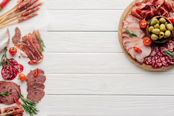 top view of arrangement of various meat snacks and olives on white wooden backdrop - Фото, изображение
