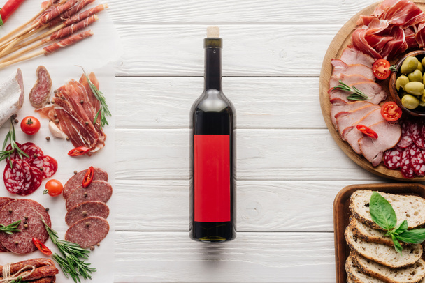 Draufsicht auf eine Flasche Rotwein und verschiedene Fleischsnacks auf einer weißen Holzplatte - Foto, Bild