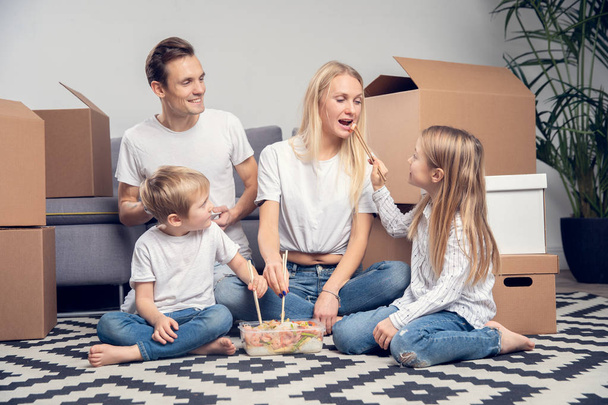 Photo of young couple with children eating rice with shrimps sitting on floor among cardboard boxes - Photo, Image