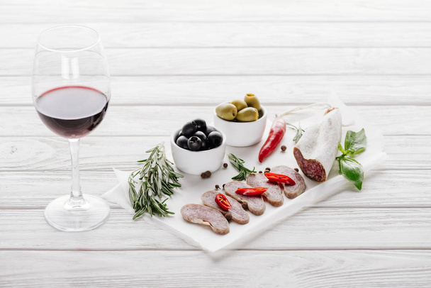close up view of glass of red wine, olives and assorted meat snacks on white wooden tabletop - Фото, изображение