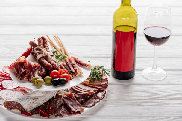Close-up beeld van fles en glas rode wijn en diverse vlees voorgerechten met olijven op witte houten tafelblad - Foto, afbeelding