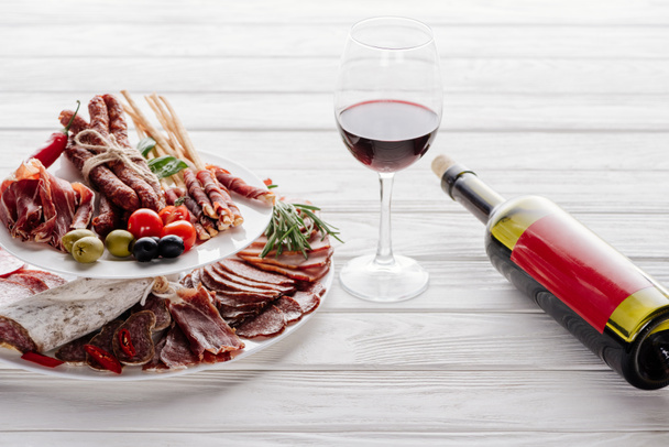 close up view of bottle and glass of red wine and various meat appetisers with olives on white wooden tabletop - Photo, Image