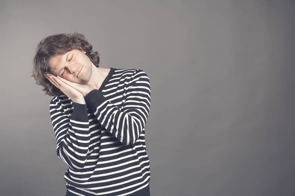 Young man in striped black and white sweater wants to sleep. Put his head in his hands and closed eyes while smiling. Standing on the grey background. Hands under his cheek. Insomnia concept. - Foto, Imagem