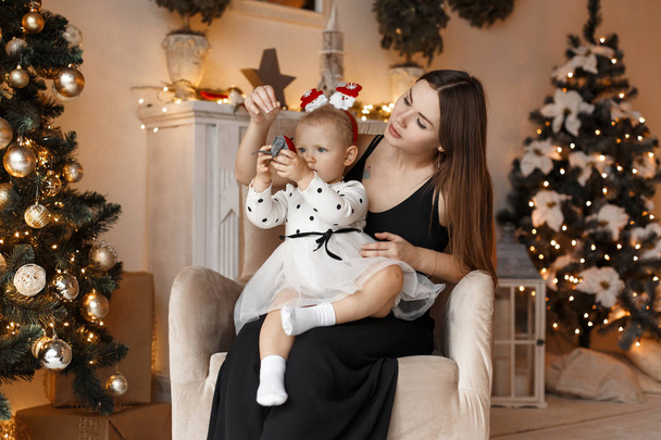 Gyönyörű bájos fiatal nő egy aranyos baba szép elegáns divatos ruhák, ül egy széken, egy újévi ünnepi szobában világos koszorúkat. Boldog karácsonyi családi ünnepek - Fotó, kép