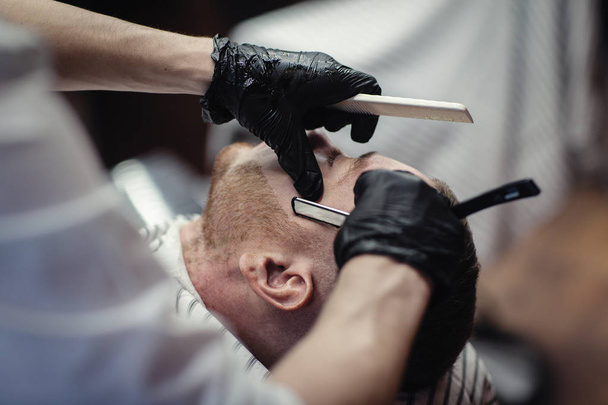 Hairdresser in black gloves shaves the razor's beard of the client - Fotó, kép